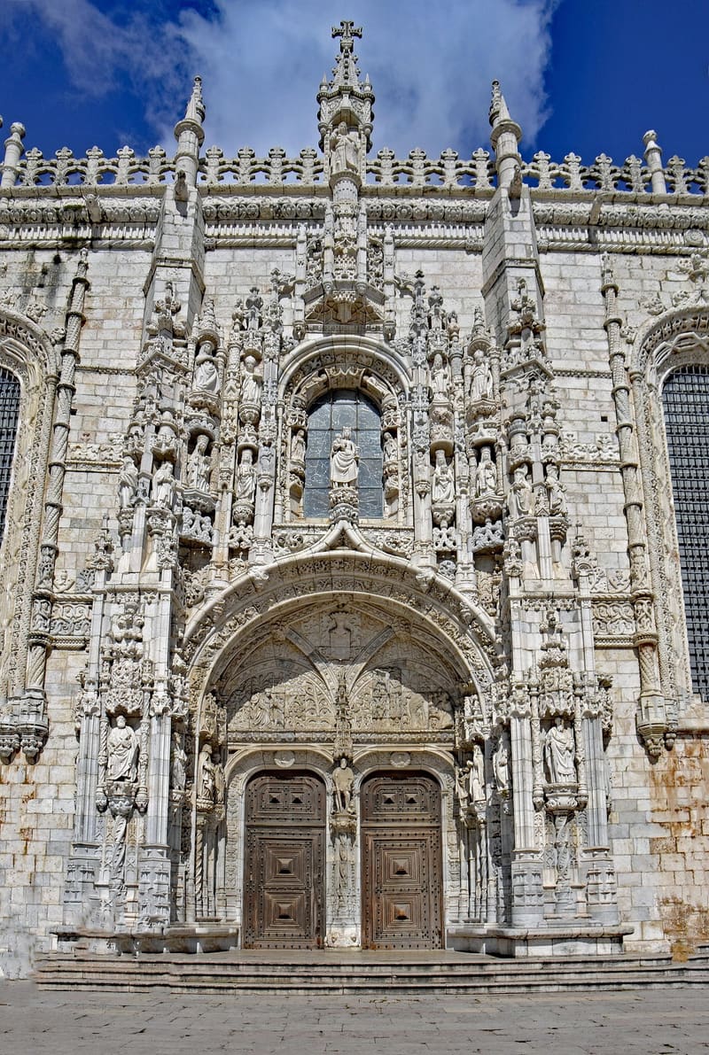 Mosteiro dos Jeronimos in Belem, Lisbon — visitors crossing Praca do Imperio along the long Manueline limestone facade with its lantern dome and ornate carved south portal. UNESCO World Heritage Site since 1983, the resting place of Vasco da Gama and Luis de Camoes.